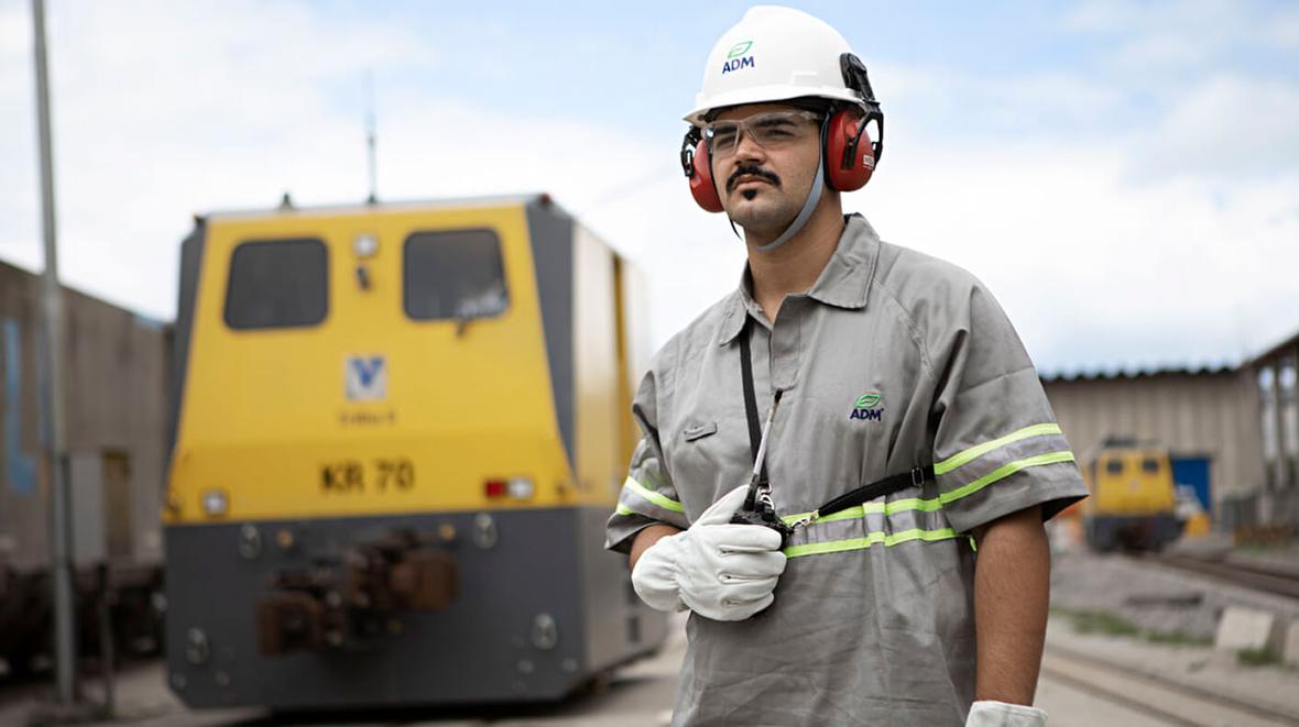 An ADM employee wearing a hardhat, standing in a train yard and holding a walkie talkie