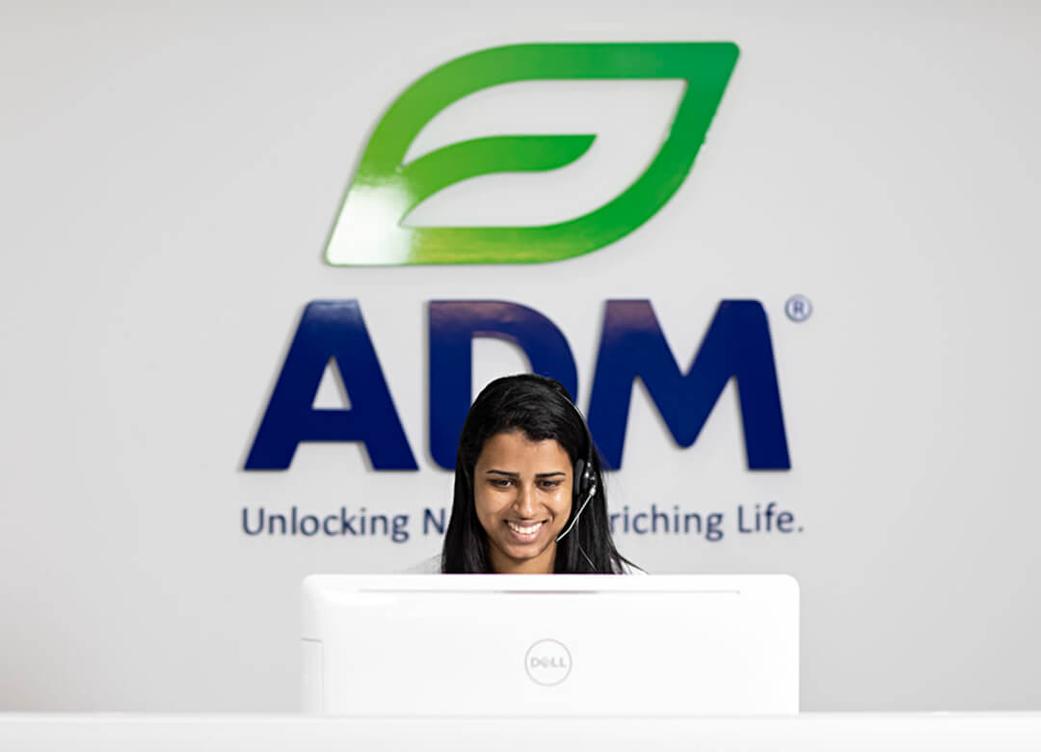 An employee wearing a headset and sitting behind a computer with a large ADM logo on the wall behind them