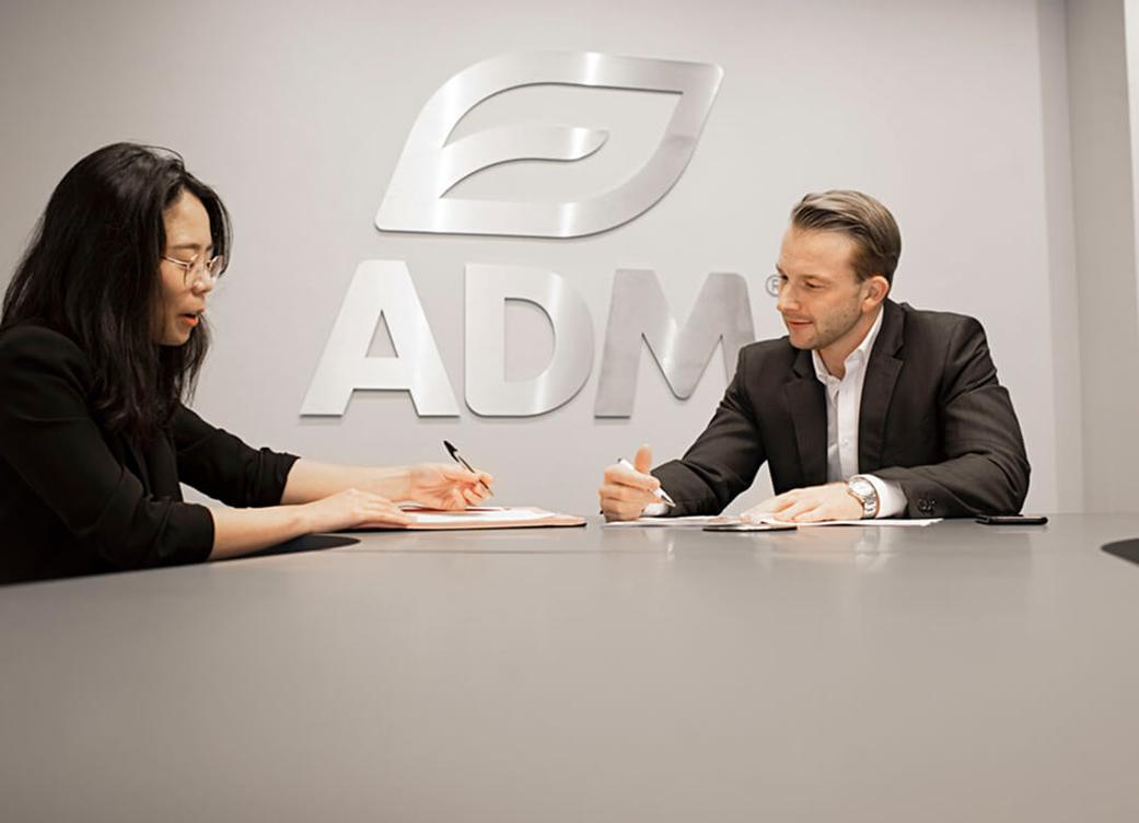 Two ADM employees sitting at a table and writing with a large ADM logo on the wall behind them