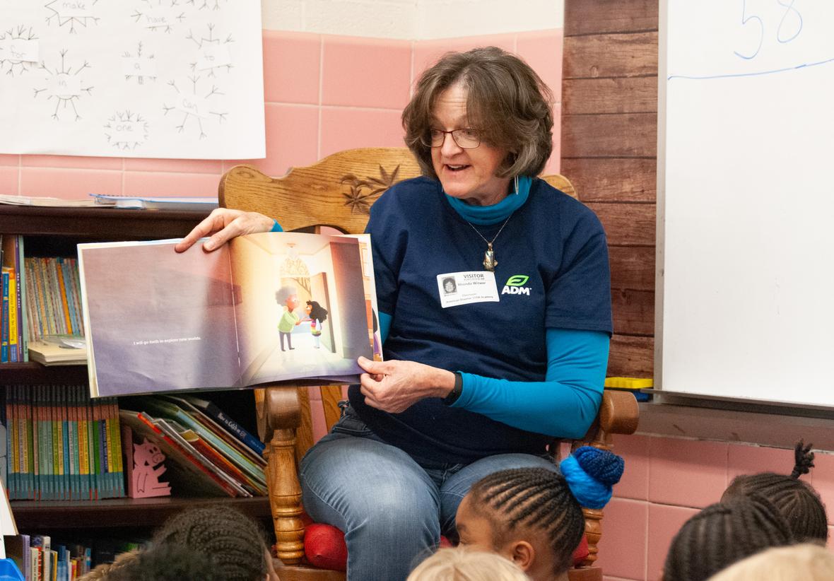 A woman in a classroom, holding up a picture book and reading out loud.