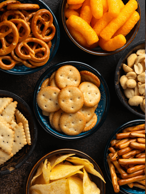 Multiple bowls of snacks including pretzels, cheese curls, cracker, potato chips, and nuts