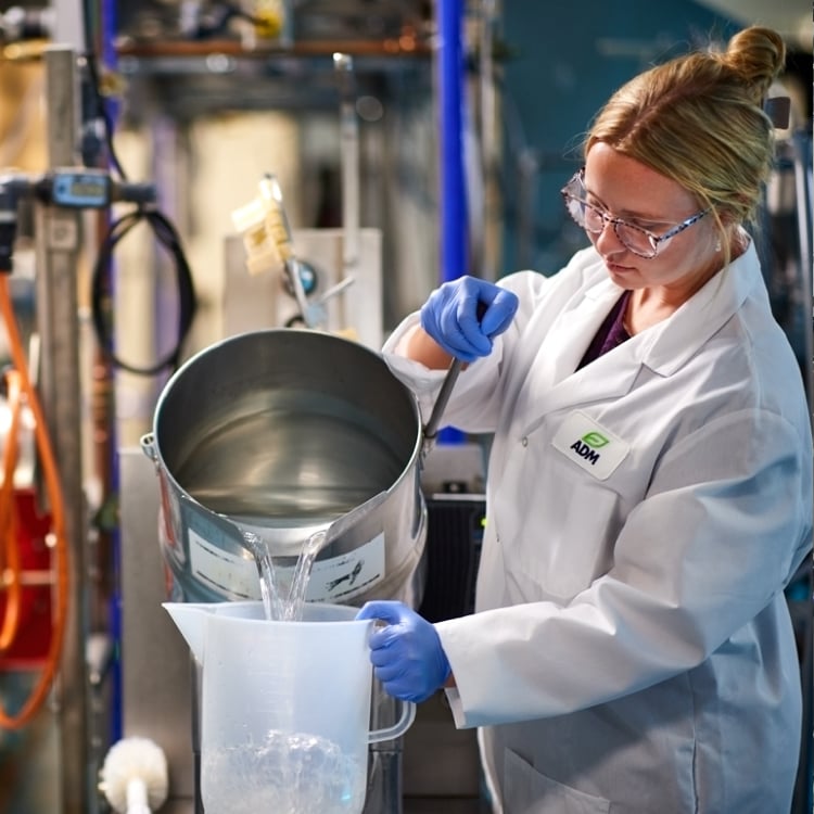 A scientist in a white ADM lab coat pouring liquid from a metal pot into a pitcher