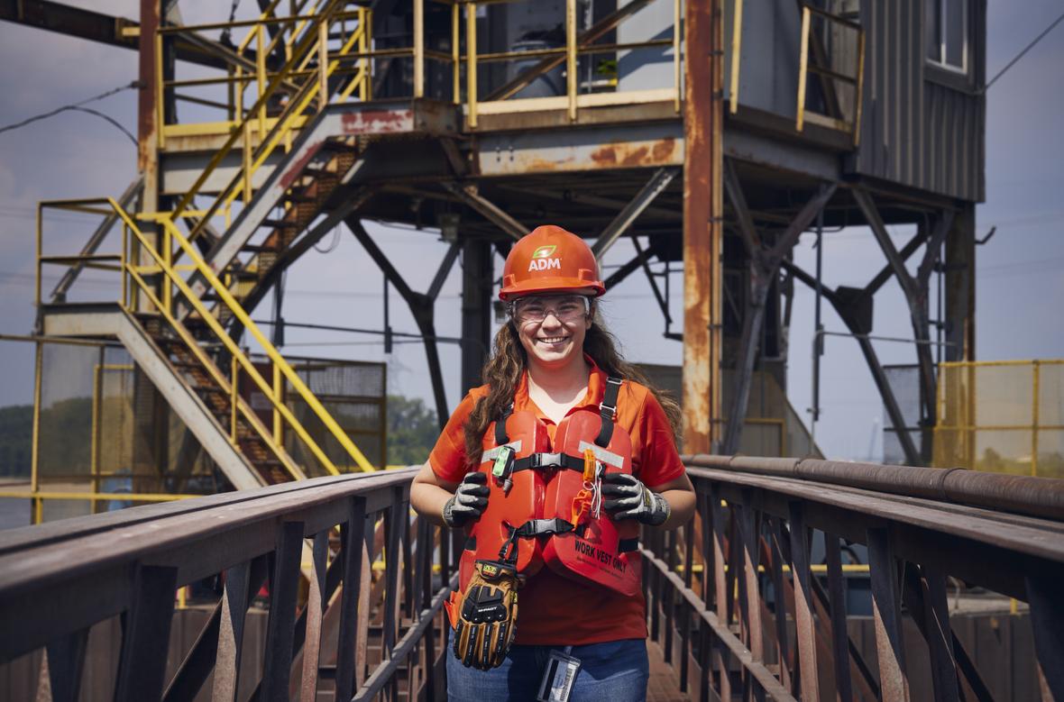 Female worker smiling wearing safety gear and helmet with ADM logo on bridge.