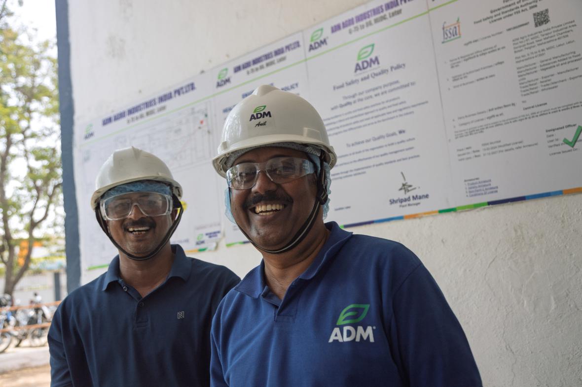 Two smiling ADM employees at an India facility wear white hard hats, safety glasses, and hairnets, standing in front of a facility diagram poster in the background.