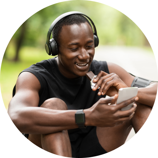 A man sitting on the ground wearing headphones and looking at his phone while eating a protein bar.