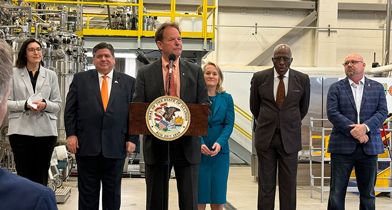 A group of people wearing suits in a factory setting. A man is speaking behind a lectern.